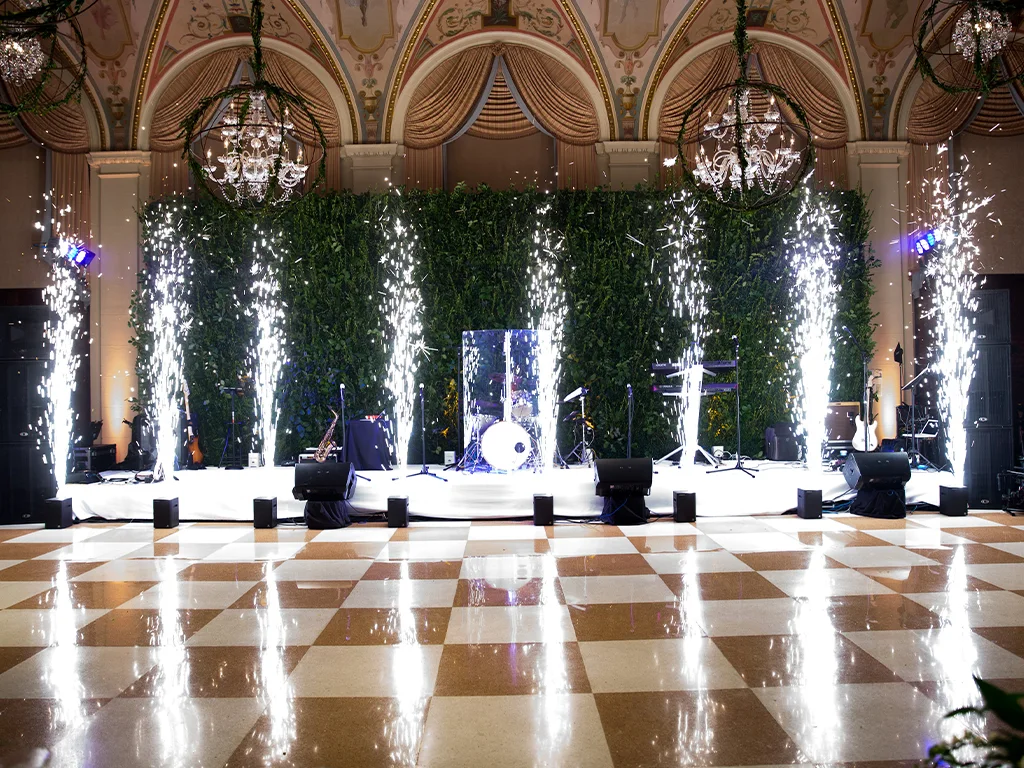 Live band stage with drum kit, guitars and microphones against a green wall backdrop, cold-spark fountains firing beside a checkered dance floor under hanging chandeliers.