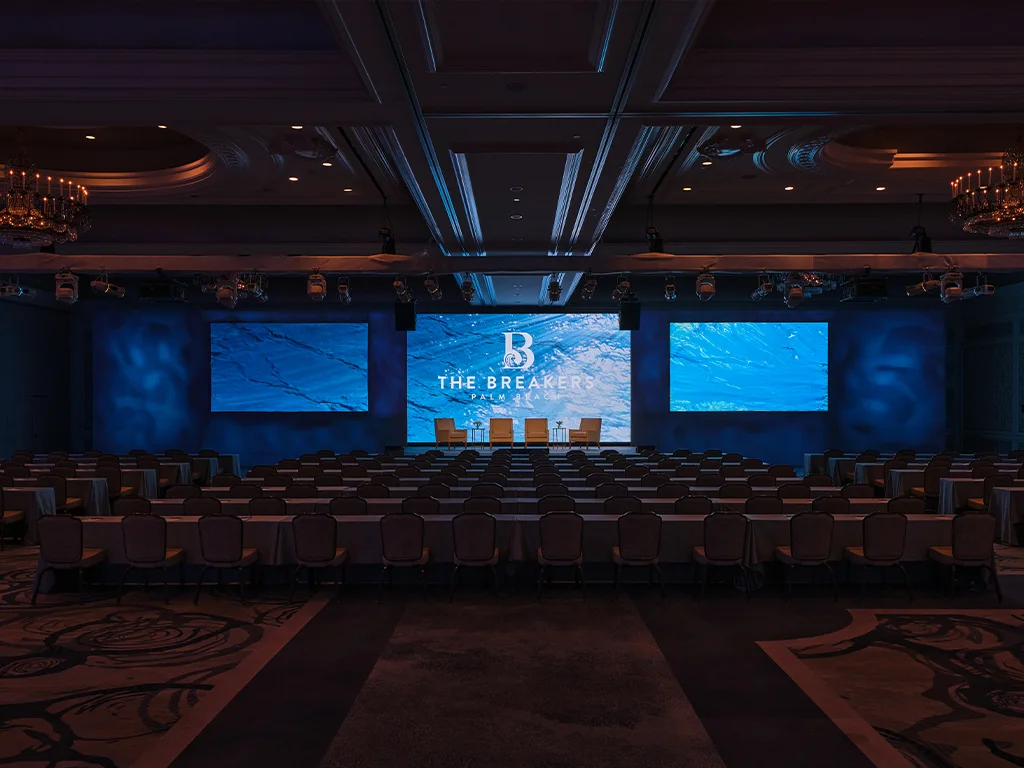 Three large LED screens on a lit stage displaying wave imagery and The Breakers Palm Beach logo behind a panel setup of four chairs, facing rows of skirted tables under a truss of spotlights.