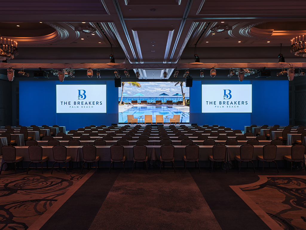 Ballroom stage with three connected screens showing The Breakers Palm Beach logo flanking a poolside image, four chairs staged in front, rows of skirted tables facing the screens under a lighting truss.