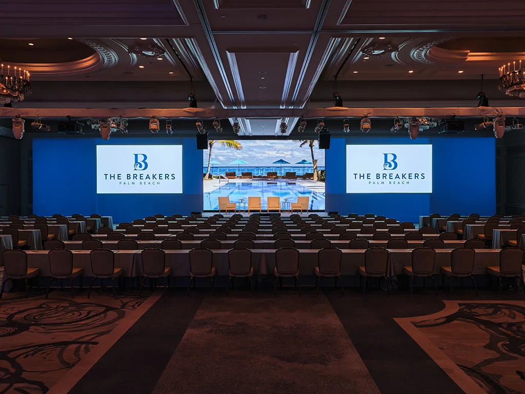 Ballroom stage with three connected screens showing The Breakers Palm Beach logo flanking a poolside image, four chairs staged in front, rows of skirted tables facing the screens under a lighting truss.