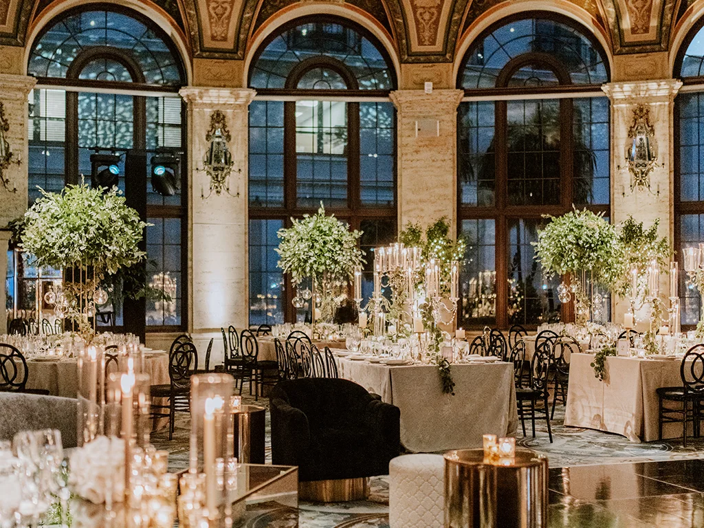 Evening reception under arched windows with tall greenery centerpieces, mixed lounge and dining seating, gold candle pillars, and stage lighting fixtures visible on a side wall.