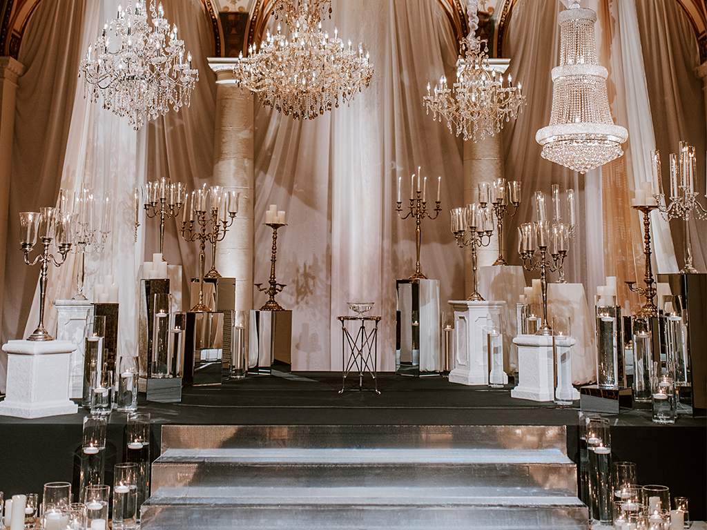 A staged display of ornate silver and crystal candelabras, pillar candles, and mirrored pedestals arranged on risers beneath four hanging crystal chandeliers against draped fabric.