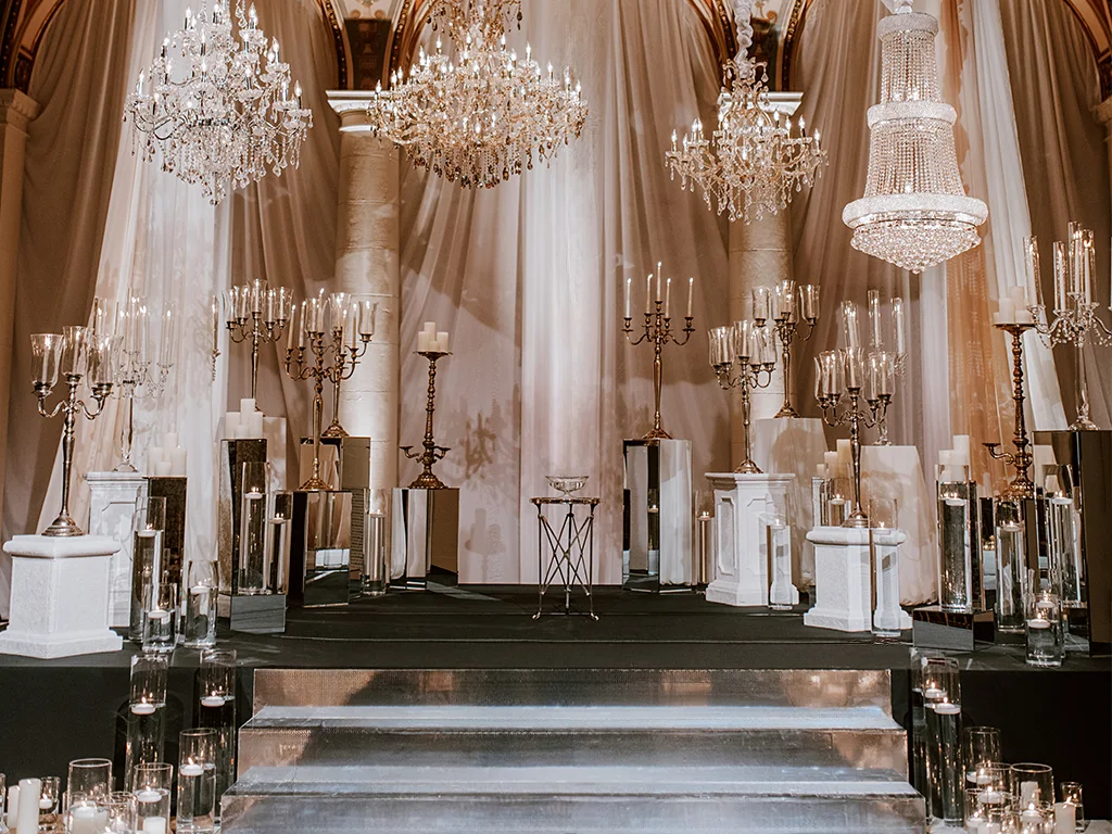 A staged display of ornate silver and crystal candelabras, pillar candles, and mirrored pedestals arranged on risers beneath four hanging crystal chandeliers against draped fabric.