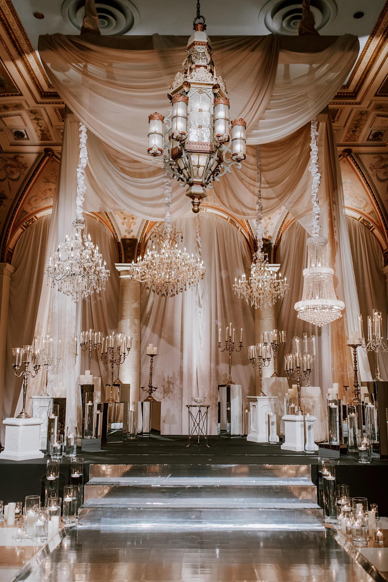 A staged display of ornate silver and crystal candelabras, pillar candles, and mirrored pedestals arranged on risers beneath four hanging crystal chandeliers against draped fabric.
