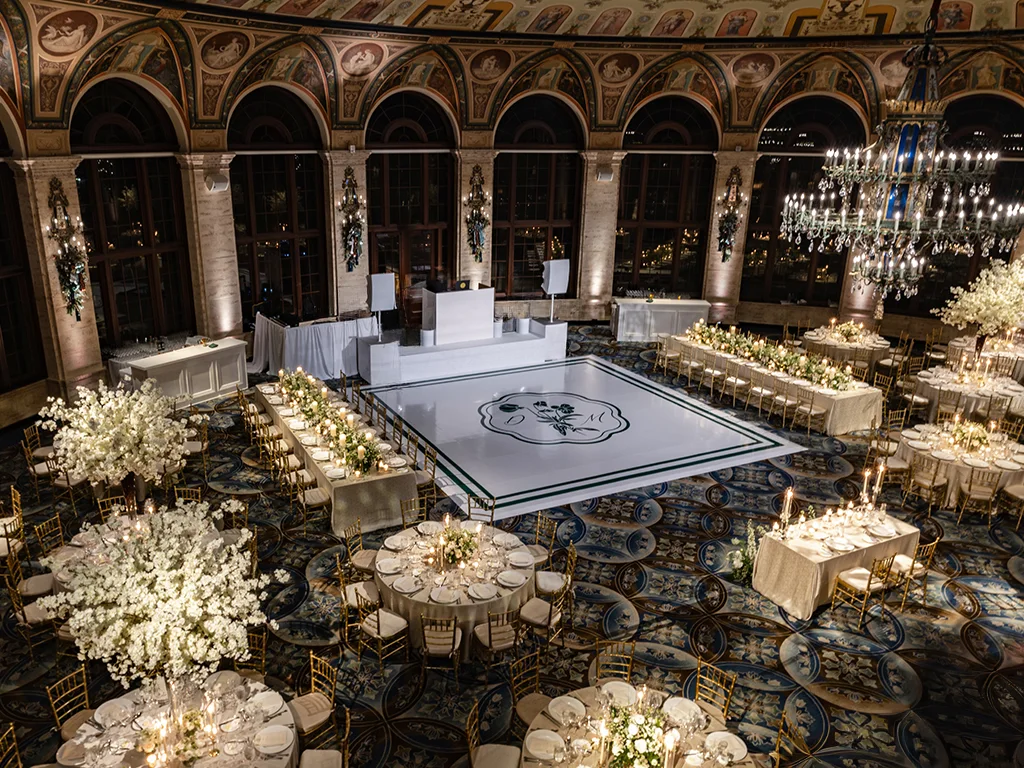 Aerial view of a frescoed ballroom with a custom monogrammed dance floor framed by round and banquet tables, white floral trees, and a DJ booth with speakers along the back wall.