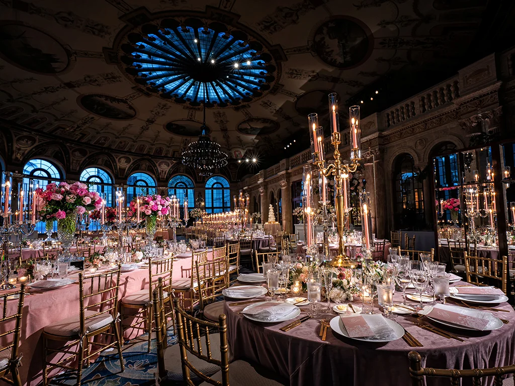 Ballroom under a stained-glass domed ceiling, set with pink and mauve linens, gold candelabras, tall tapers, and pink rose centerpieces on long and round tables.