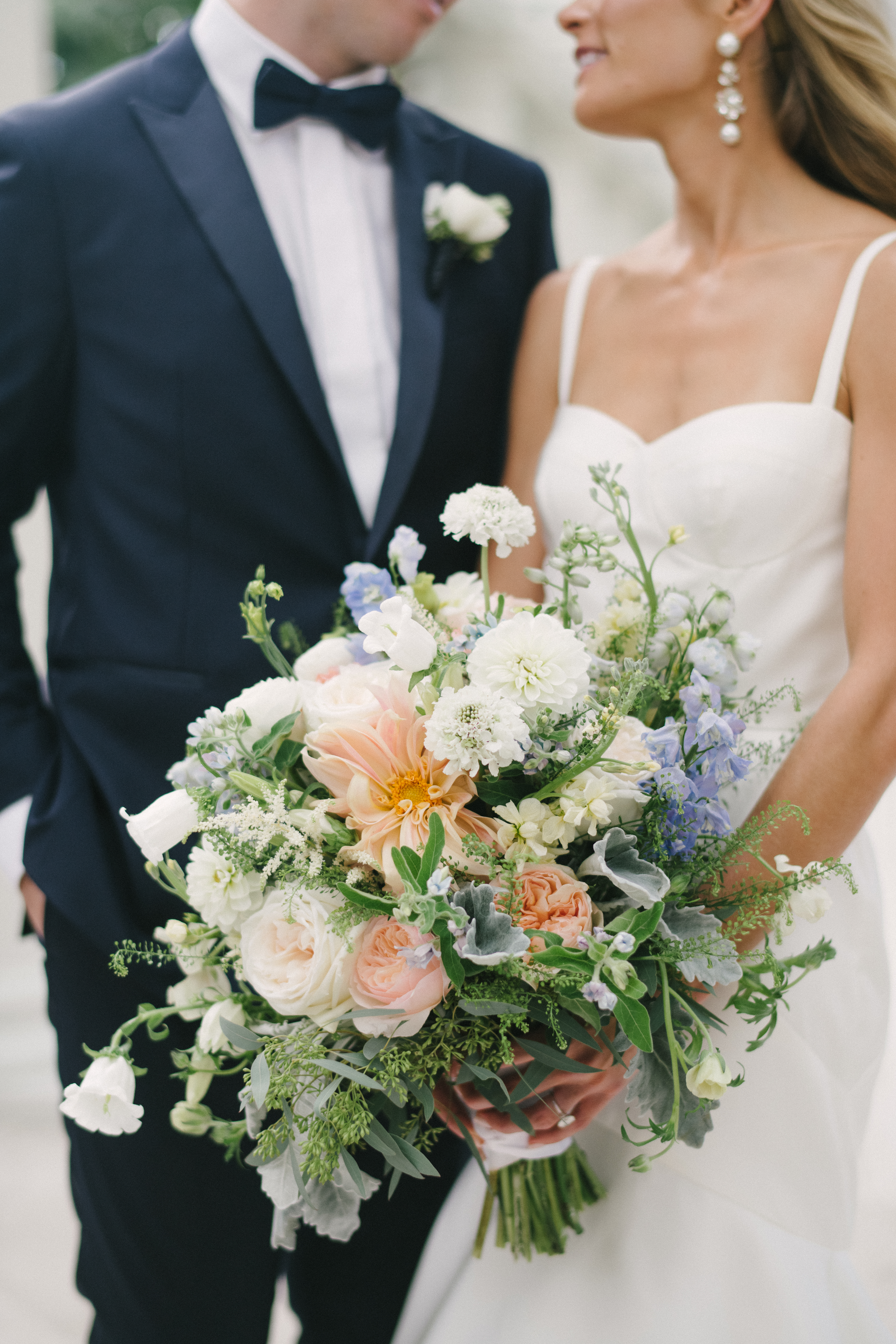 Close-up of a bridal bouquet mixing peach dahlias, blush garden roses, blue delphinium and white blooms, held between a bride in white and a groom in a navy suit.