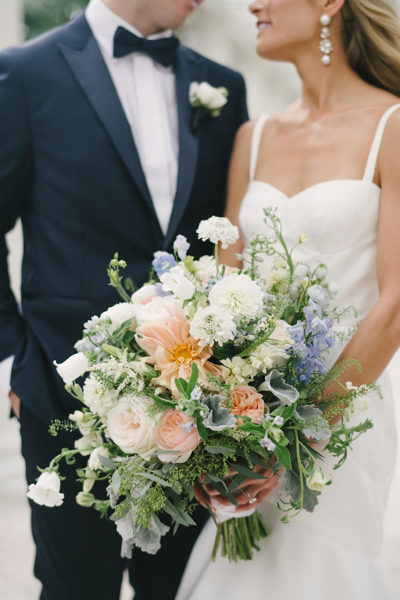 Close-up of a bridal bouquet mixing peach dahlias, blush garden roses, blue delphinium and white blooms, held between a bride in white and a groom in a navy suit.