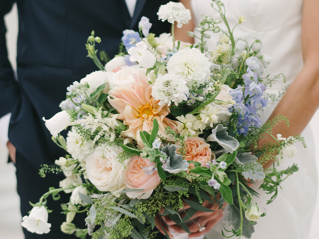 Close-up of a bridal bouquet mixing peach dahlias, blush garden roses, blue delphinium and white blooms, held between a bride in white and a groom in a navy suit.