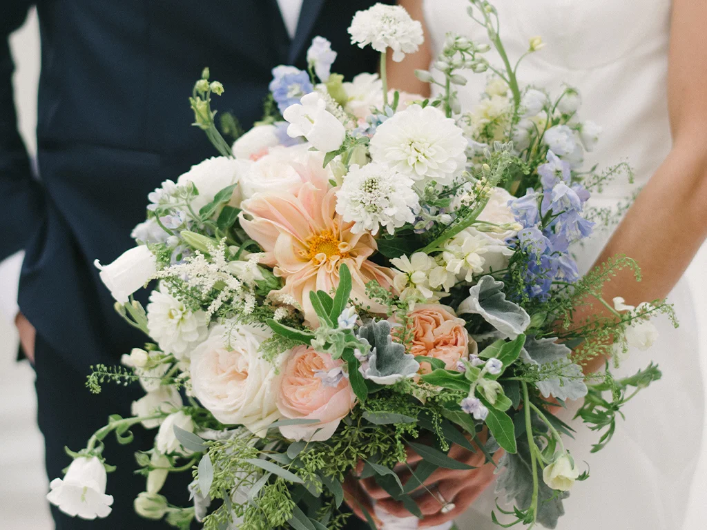 Close-up of a bridal bouquet mixing peach dahlias, blush garden roses, blue delphinium and white blooms, held between a bride in white and a groom in a navy suit.