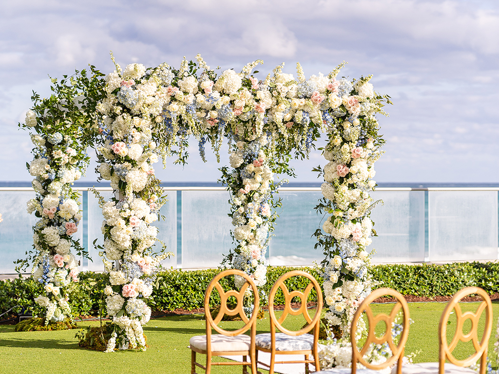 Four-post ceremony chuppah covered in white hydrangea and blush roses on an oceanfront lawn, with gold ring-back chairs and the sea beyond a glass railing.