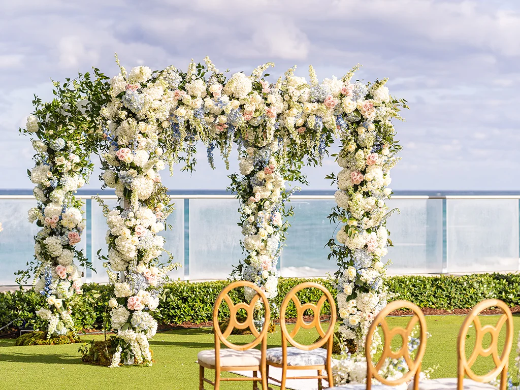 Four-post ceremony chuppah covered in white hydrangea and blush roses on an oceanfront lawn, with gold ring-back chairs and the sea beyond a glass railing.