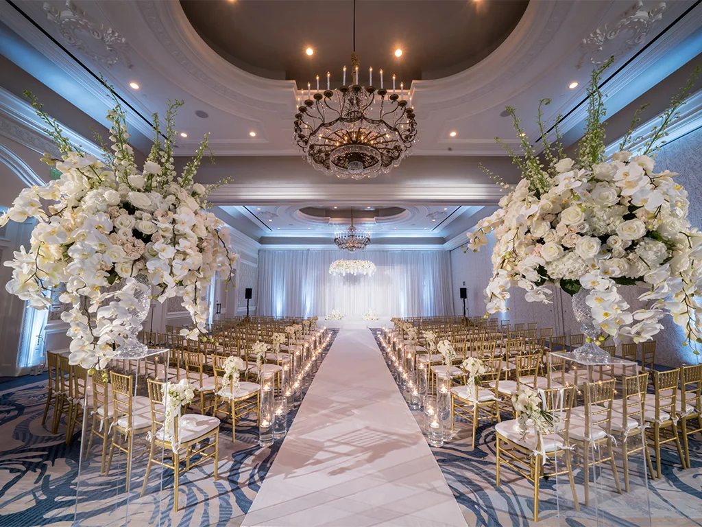 Ballroom ceremony aisle lit in blue, flanked by tall white orchid and rose pedestal arrangements and gold chairs, leading to a draped stage beneath a crystal chandelier.