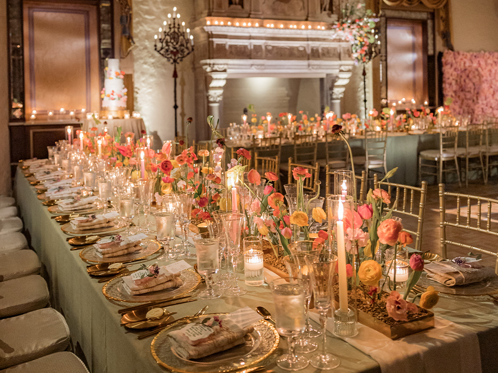 Long banquet table set with gold chargers, taper candles and low troughs of mixed pink and orange ranunculus, with a pink flower wall and iced cake visible behind.