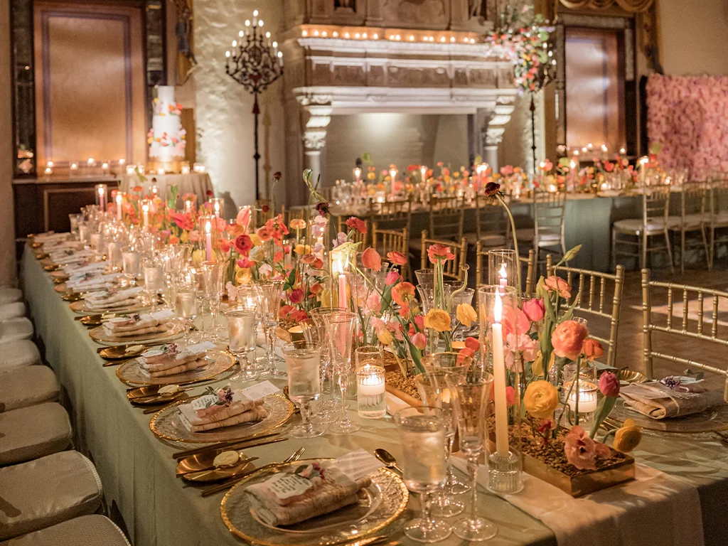 Long banquet table set with gold chargers, taper candles and low troughs of mixed pink and orange ranunculus, with a pink flower wall and iced cake visible behind.