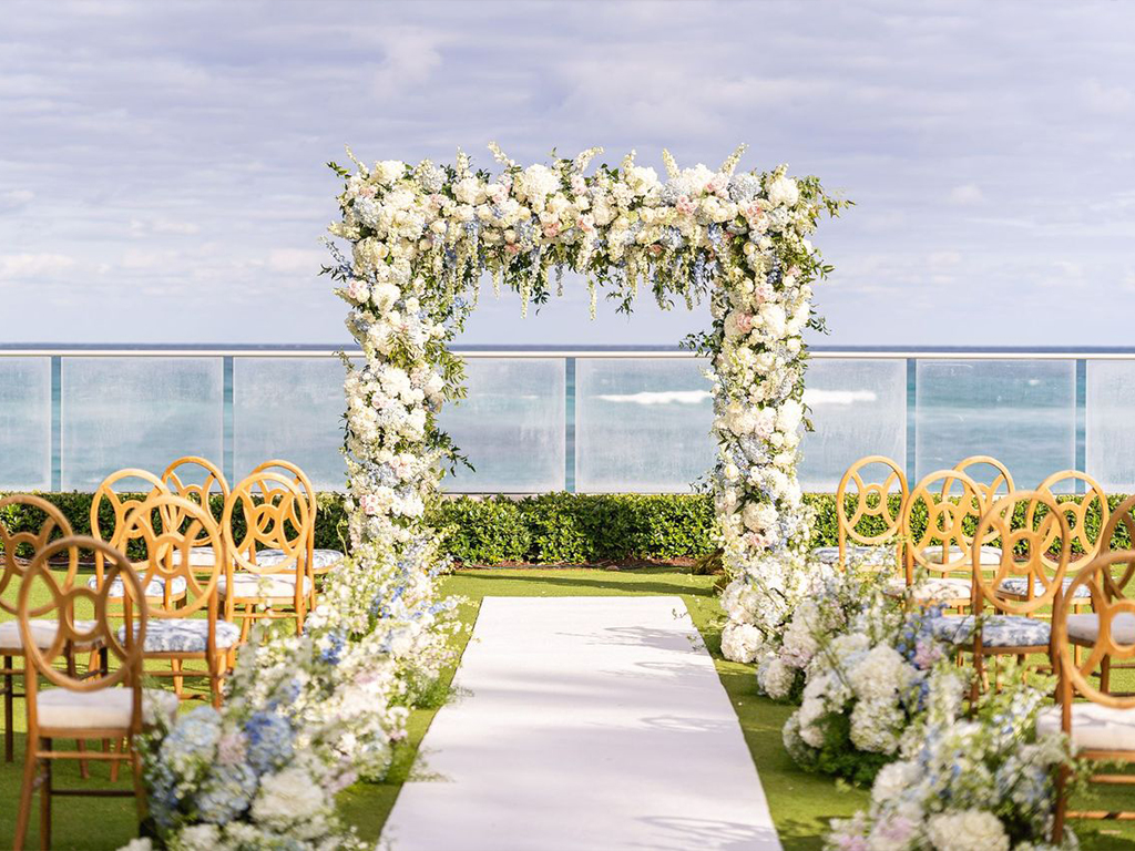 Oceanfront ceremony arch of white and blue florals over a white aisle runner, with gold ring-back chairs and matching floral clusters on the lawn, sea visible beyond the railing.
