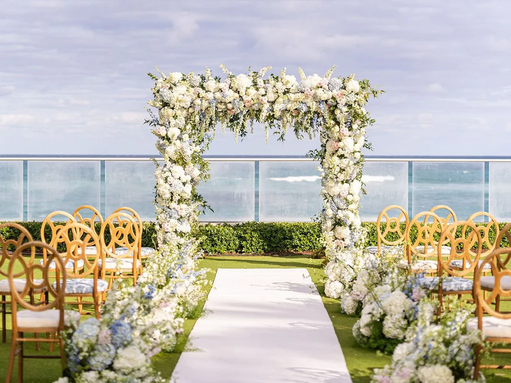 Oceanfront ceremony arch of white and blue florals over a white aisle runner, with gold ring-back chairs and matching floral clusters on the lawn, sea visible beyond the railing.