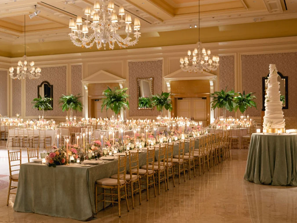Ballroom set with a long banquet table in sage linens, tall fern arrangements, candlelight, crystal chandeliers, and a tiered white wedding cake.
