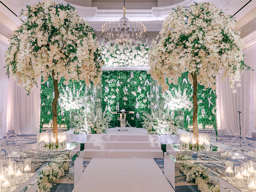 Ceremony stage with two flowering trees framing a greenery wall backdrop, a rose-draped chandelier overhead, mirrored aisle risers lined with candles, and a microphone stand at the edge of the stage.