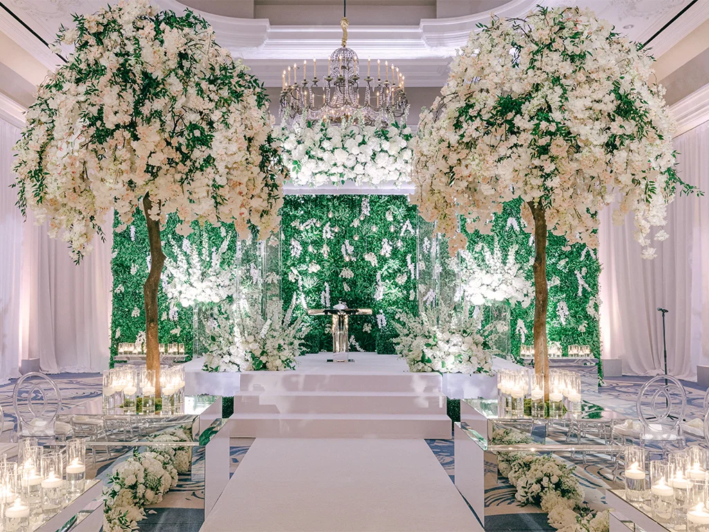 Ceremony stage with two flowering trees framing a greenery wall backdrop, a rose-draped chandelier overhead, mirrored aisle risers lined with candles, and a microphone stand at the edge of the stage.