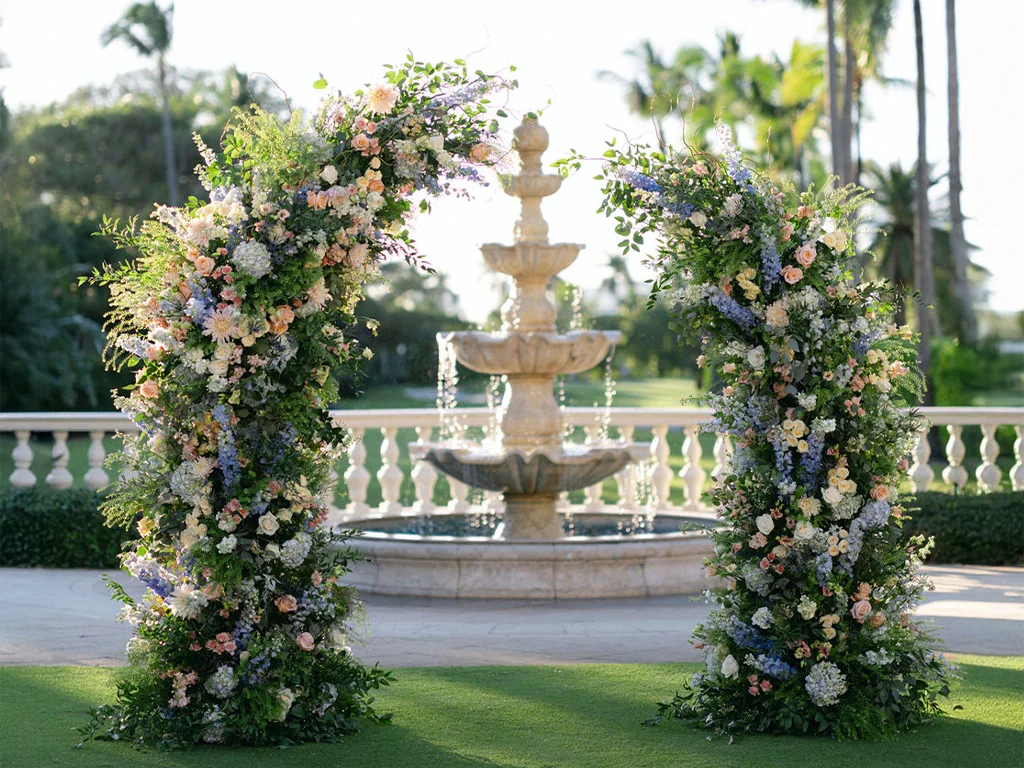 Split ceremony arch of peach, blue and white florals framing a stone tiered fountain on an outdoor terrace lined with palms.