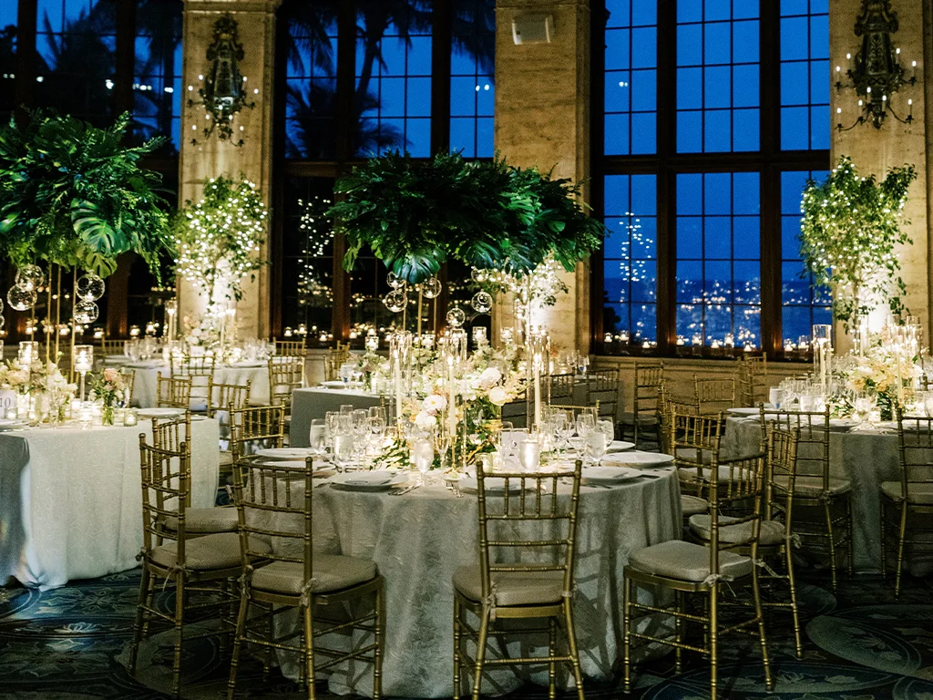 Round tables set for an evening reception with tall monstera leaf centerpieces, hanging glass orbs, candlelight and floor-to-ceiling windows overlooking the coastline at dusk.