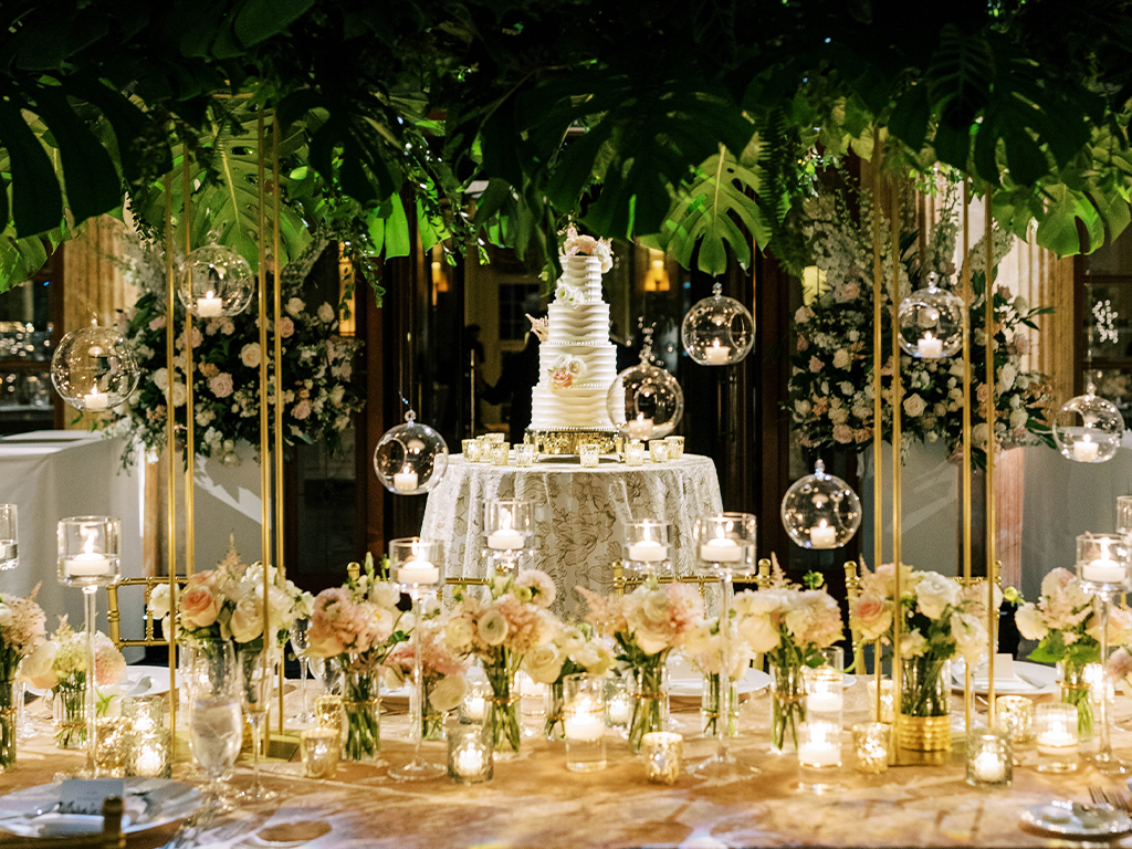 Tiered white wedding cake displayed under a monstera leaf canopy with hanging glass candle orbs and blush floral clusters lining the head table.