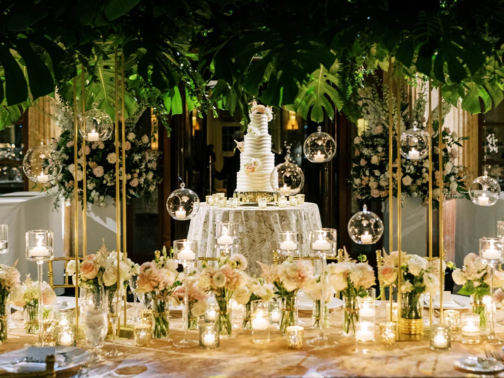Tiered white wedding cake displayed under a monstera leaf canopy with hanging glass candle orbs and blush floral clusters lining the head table.