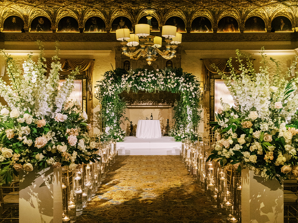 Grand indoor ceremony aisle beneath a gilded, portrait-lined ceiling, lined with candlelit floral pedestals leading to a green-and-white floral arch.