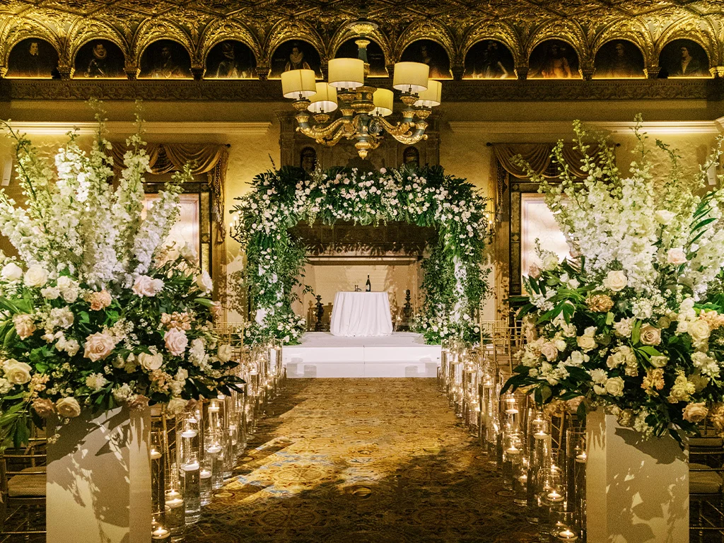 Grand indoor ceremony aisle beneath a gilded, portrait-lined ceiling, lined with candlelit floral pedestals leading to a green-and-white floral arch.