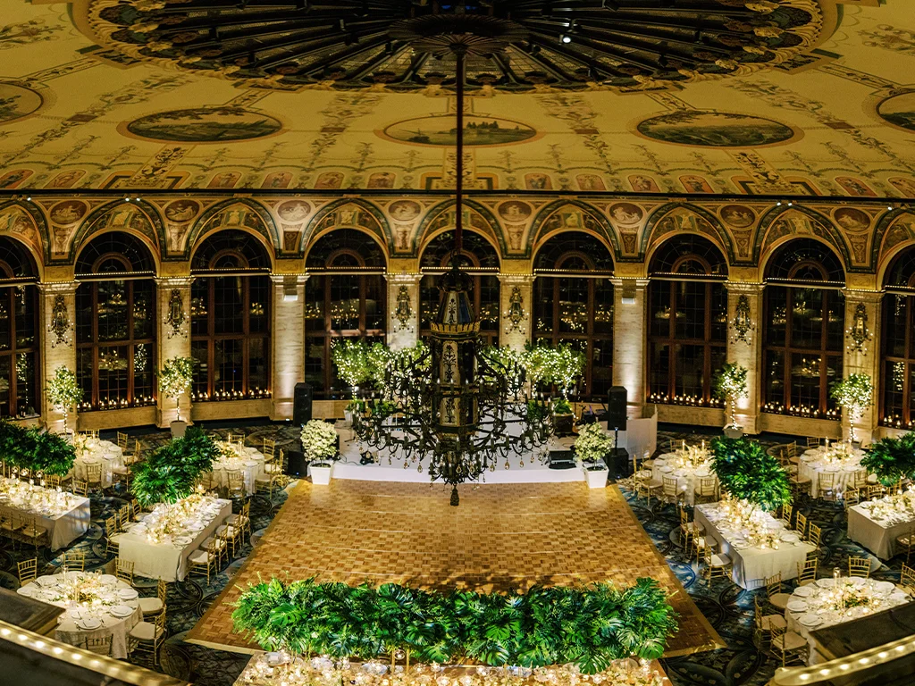 Aerial view of a domed, frescoed ballroom set for a gala with a wood dance floor framed by palm fronds, round guest tables, and speakers flanking a small stage beneath a large chandelier.