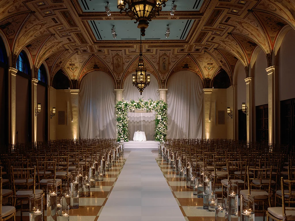 Ceremony ballroom with rows of chairs flanking a white aisle runner, floating candles, and a white floral arch against draped fabric under a painted vaulted ceiling.