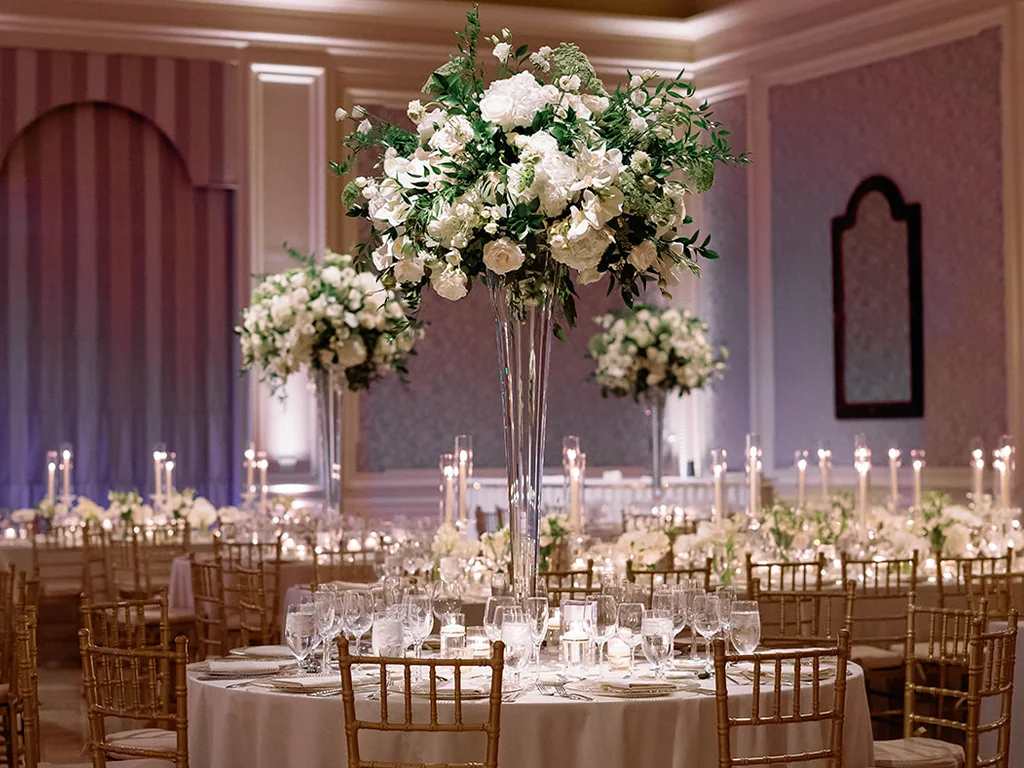 Round tables with tall trumpet-vase white rose centerpieces and rows of pillar candles, set beneath purple wall lighting.