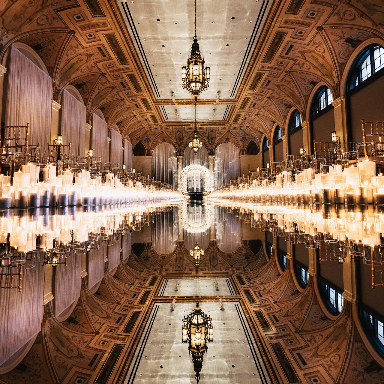 Ornate ballroom with vaulted ceilings, rows of candles lining a reflective aisle, and gold chairs.