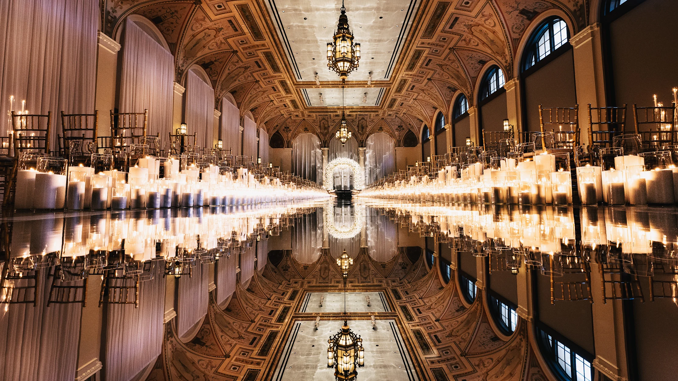 Ornate ballroom with vaulted ceilings, rows of candles lining a reflective aisle, and gold chairs.