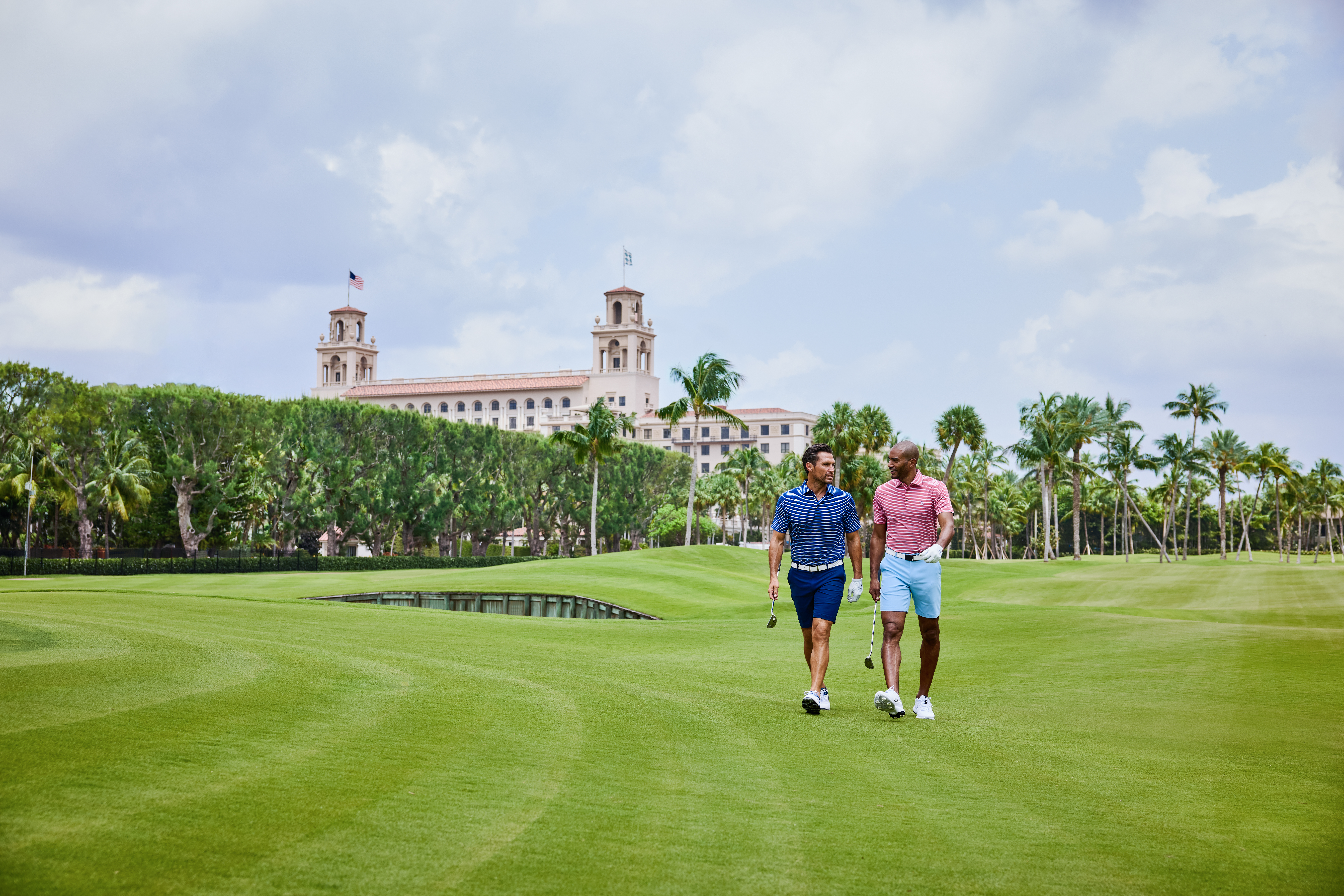 Two men walking on a lush golf course with palm trees and a Mediterranean-style resort building.