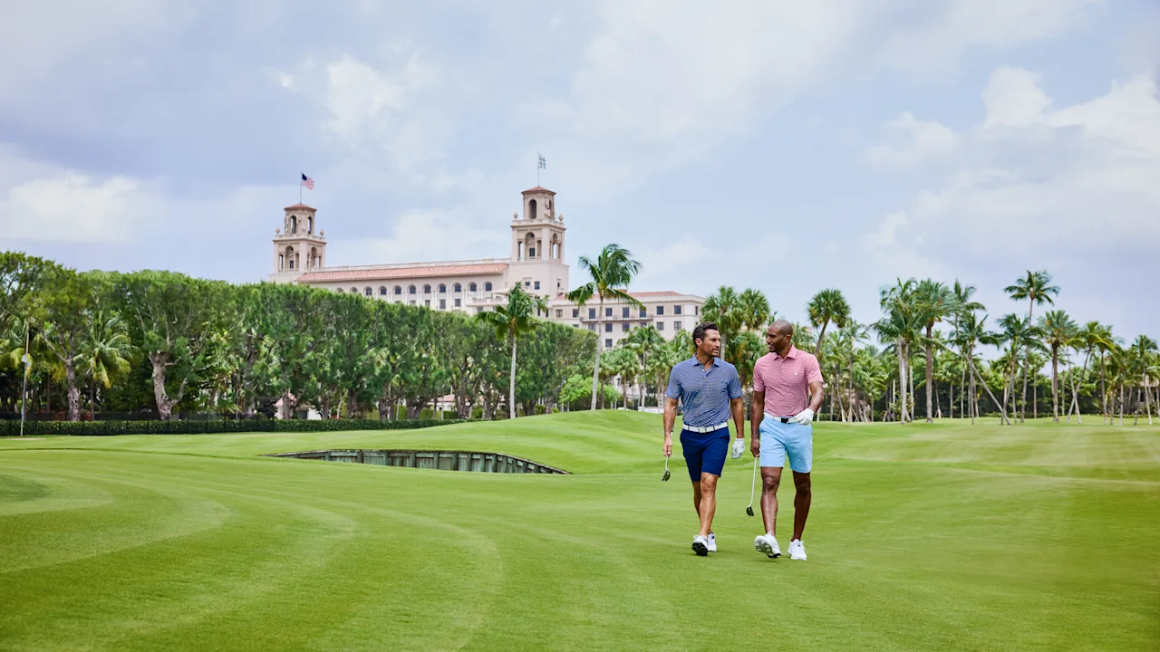 Two men walking on a lush golf course with palm trees and a Mediterranean-style resort building.