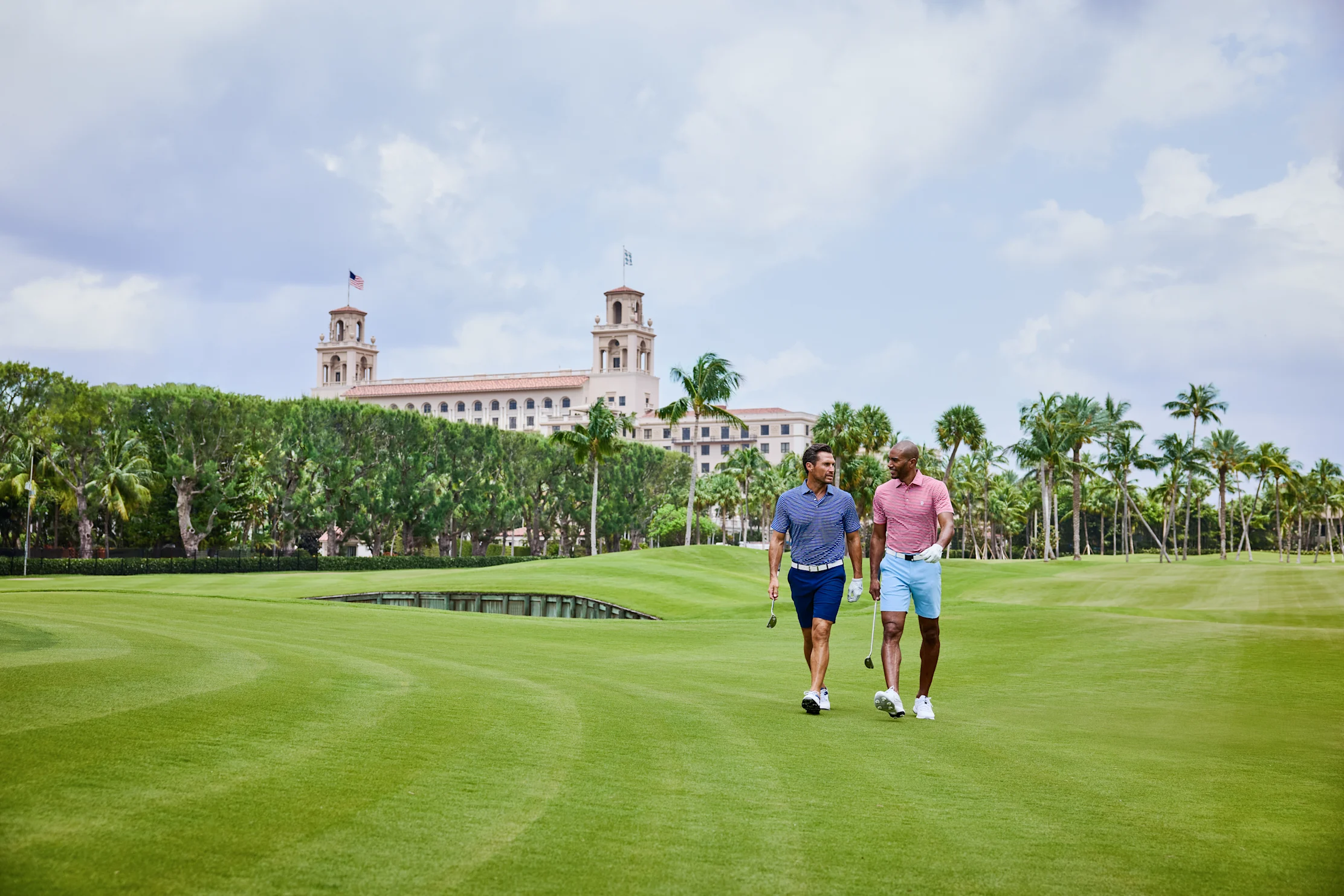 Two men walking on a lush golf course with palm trees and a Mediterranean-style resort building.