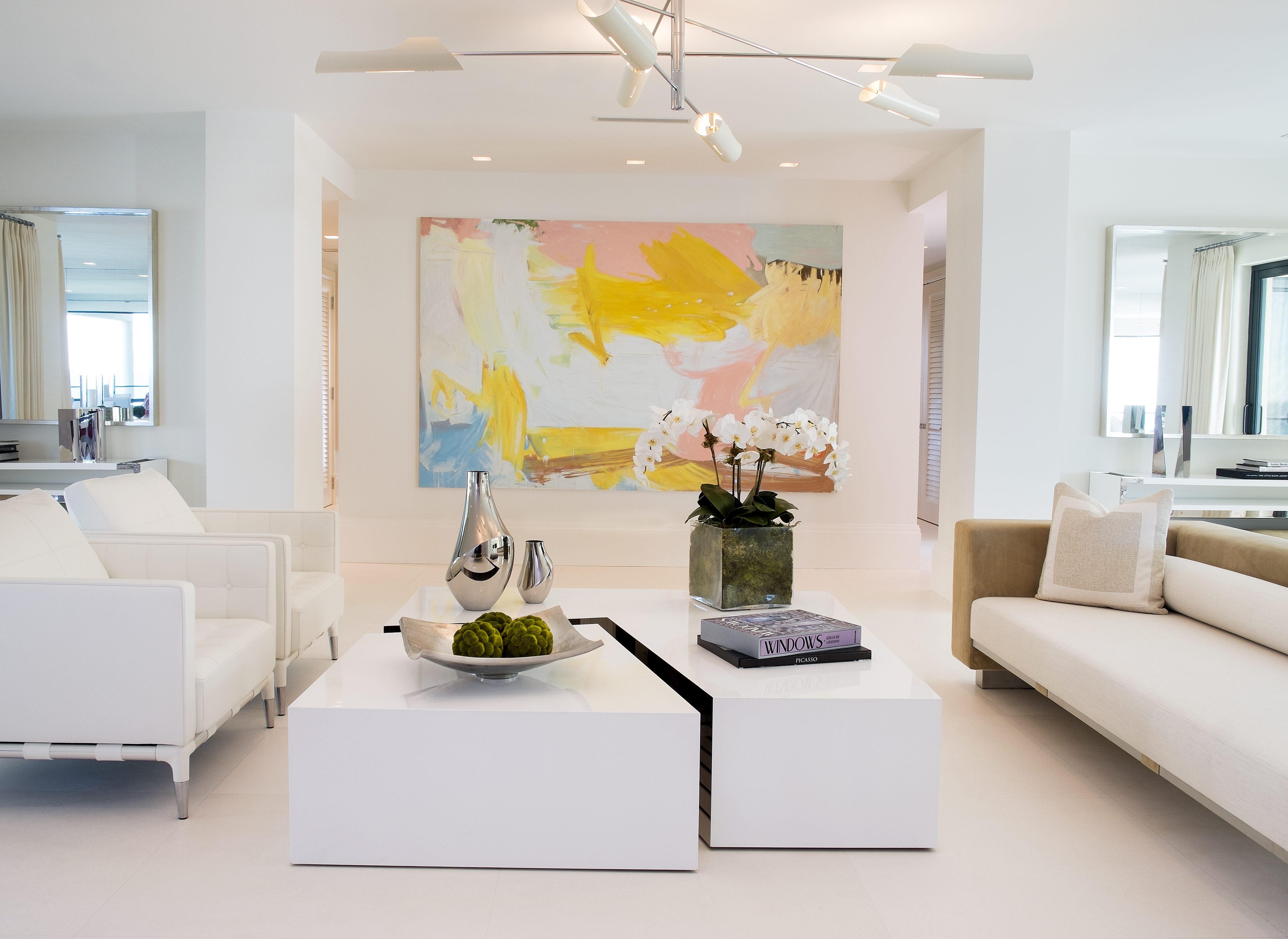 Modern white living room with colorful abstract art, white sofas, and geometric coffee tables.