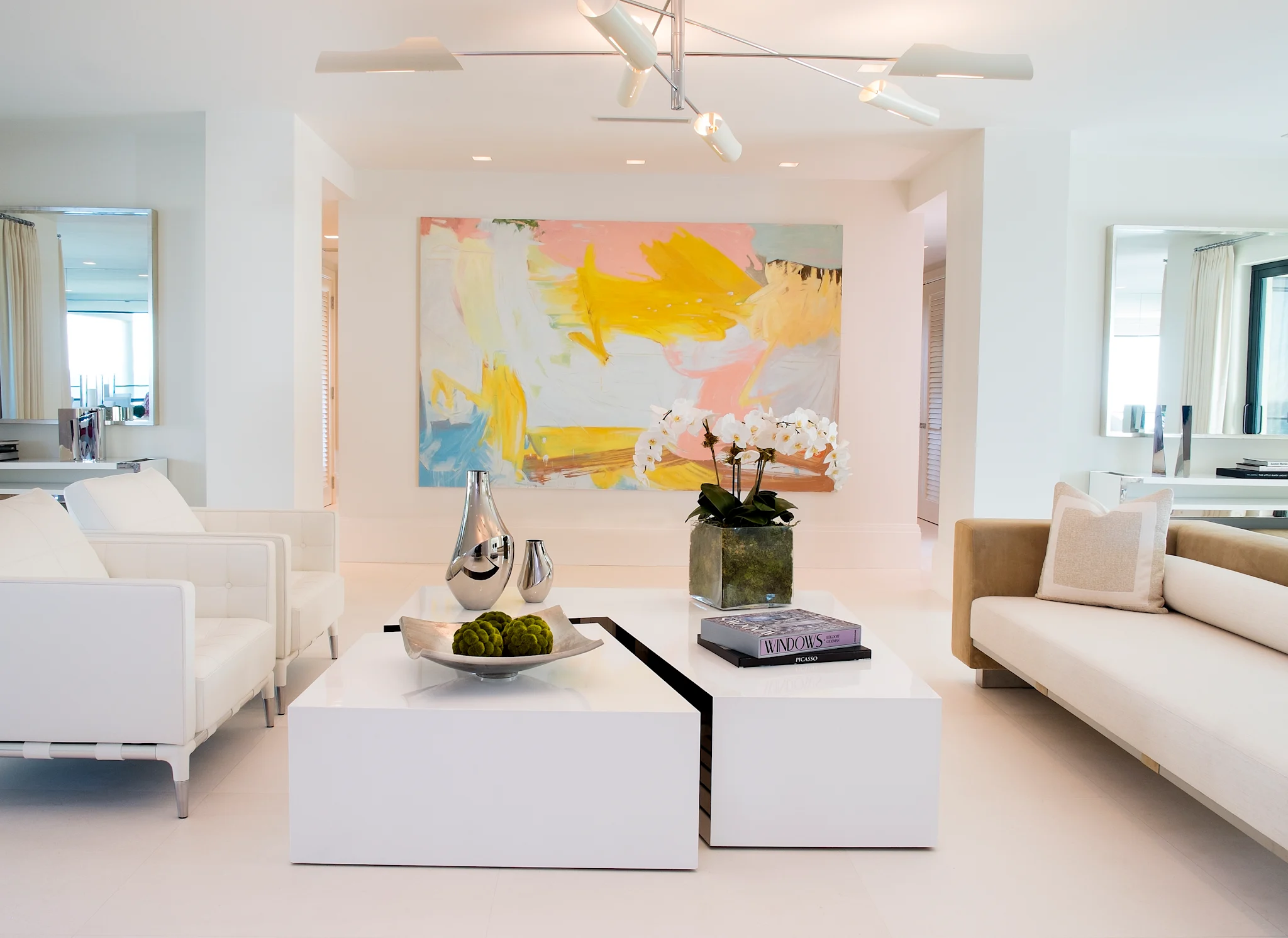 Modern white living room with colorful abstract art, white sofas, and geometric coffee tables.
