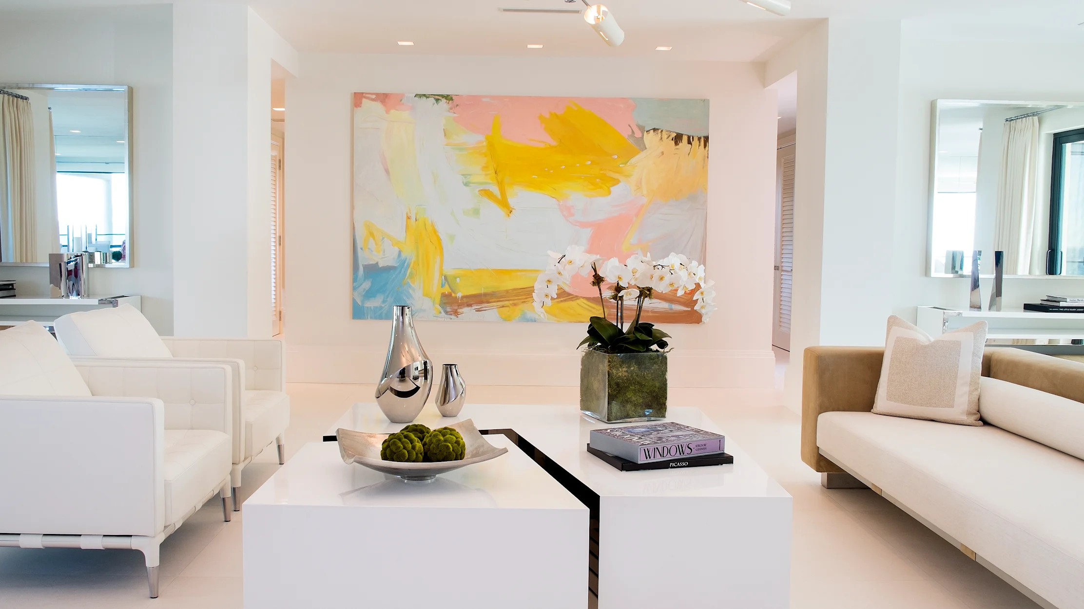 Modern white living room with colorful abstract art, white sofas, and geometric coffee tables.