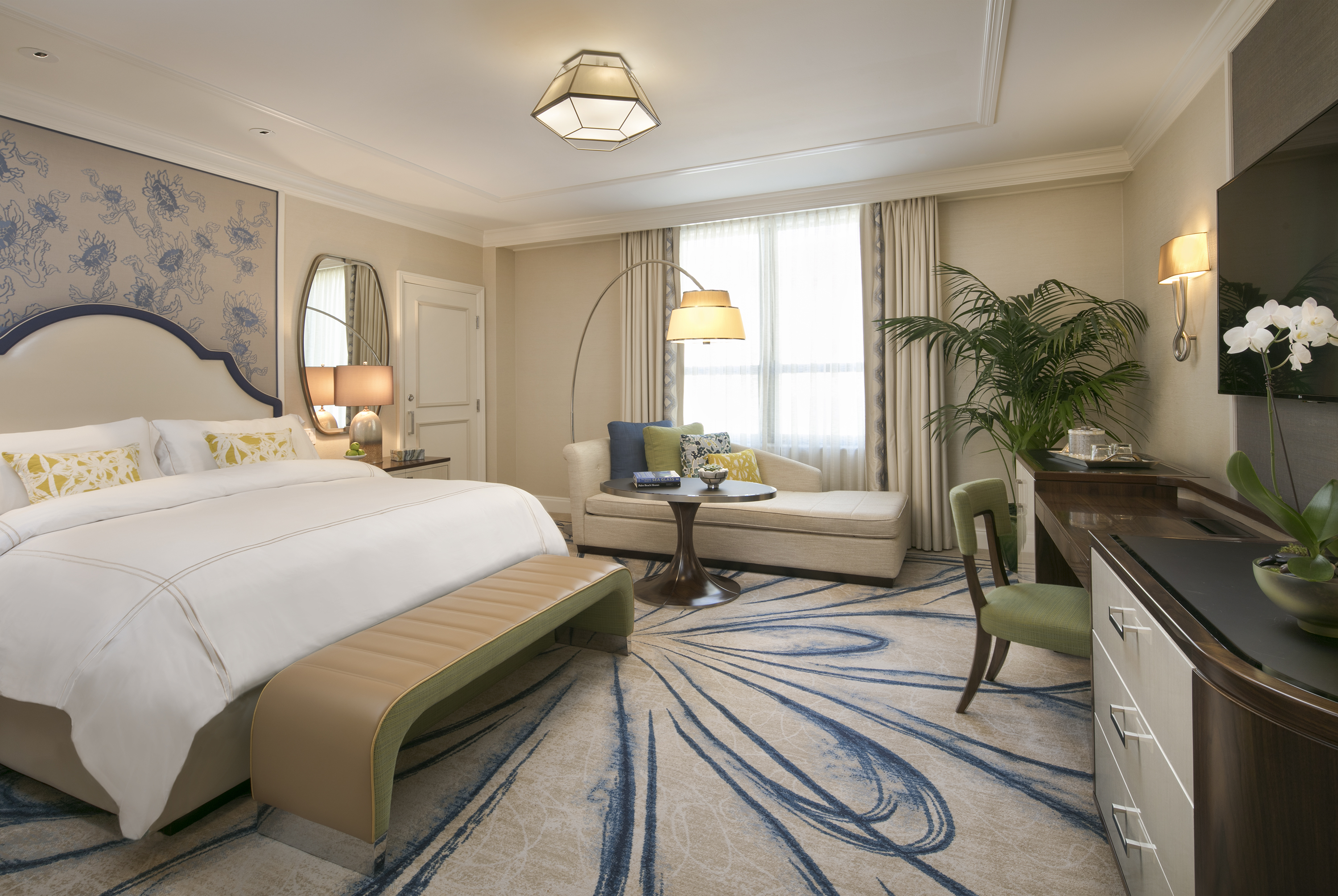 Elegant hotel bedroom with white bedding, blue headboard, chaise lounge, arc lamp, and patterned rug.