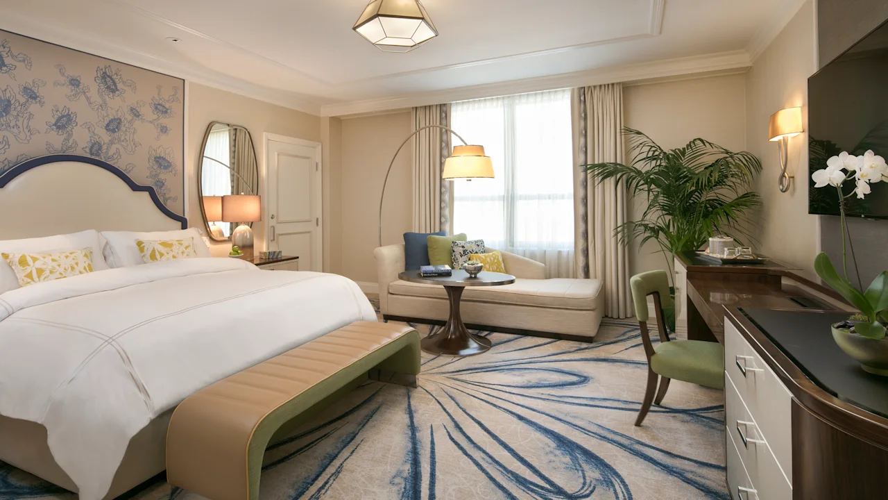 Elegant hotel bedroom with white bedding, blue headboard, chaise lounge, arc lamp, and patterned rug.