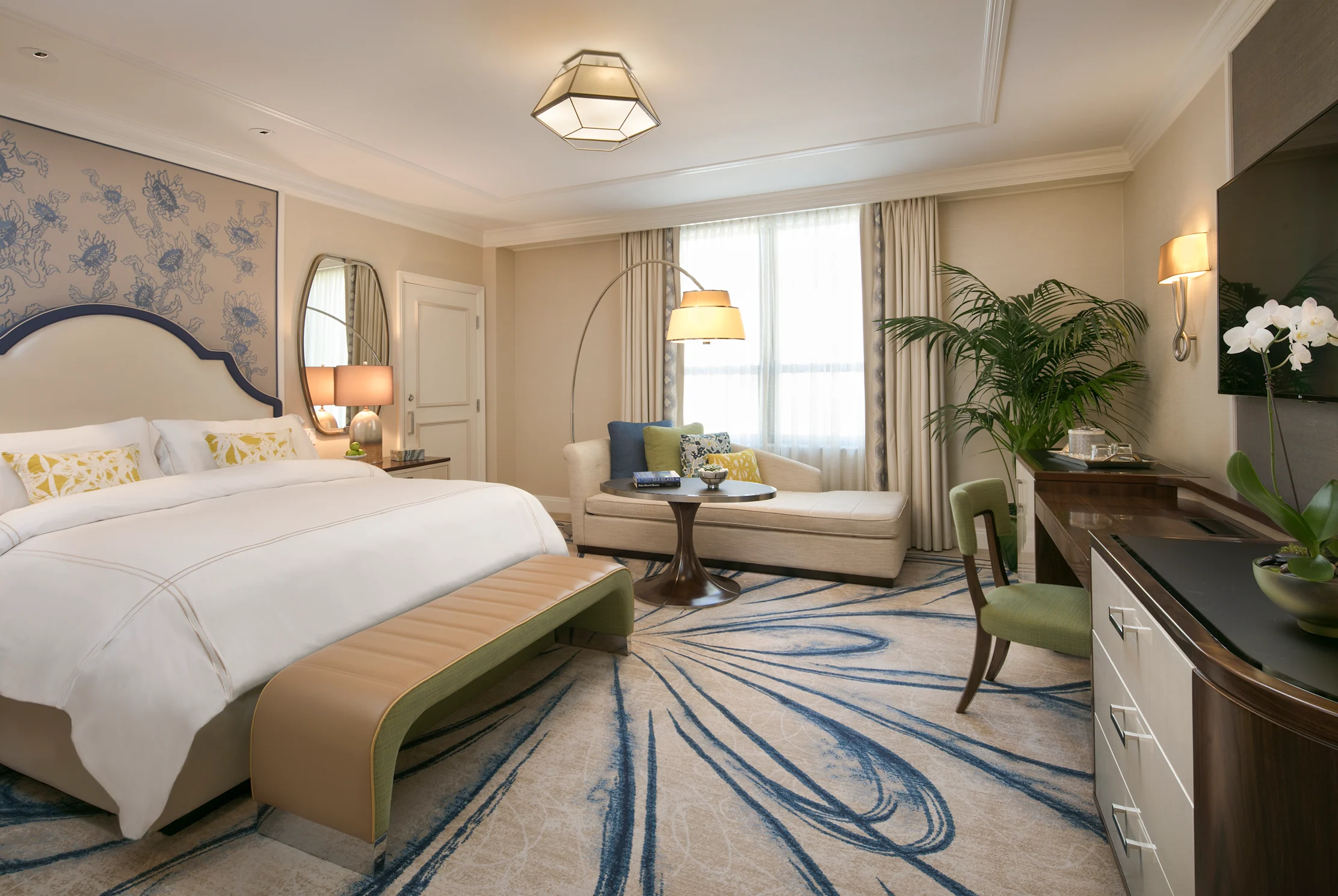 Elegant hotel bedroom with white bedding, blue headboard, chaise lounge, arc lamp, and patterned rug.
