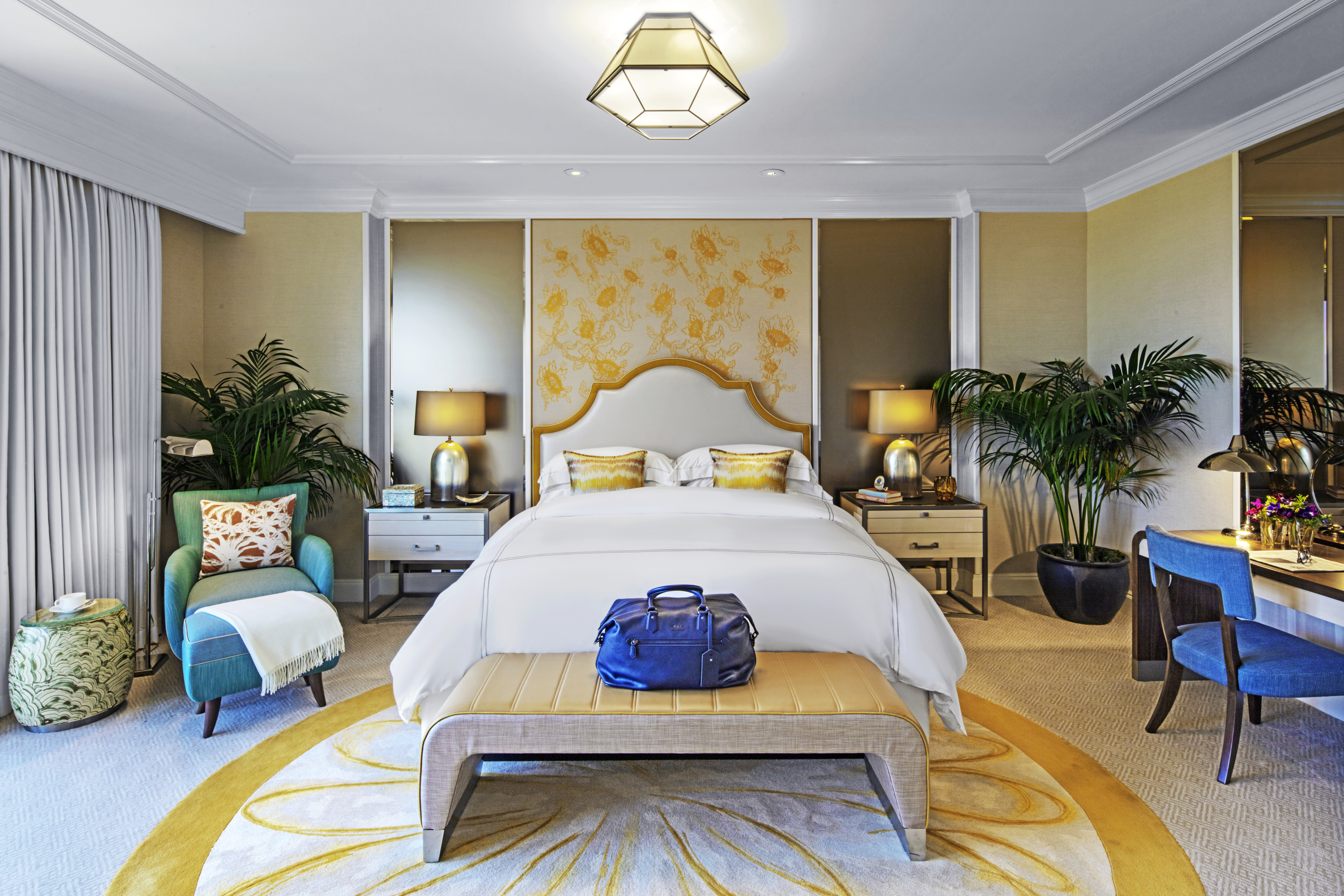 Elegant hotel bedroom with white bed, gold accents, floral wallpaper, palm plants, and geometric ceiling light.