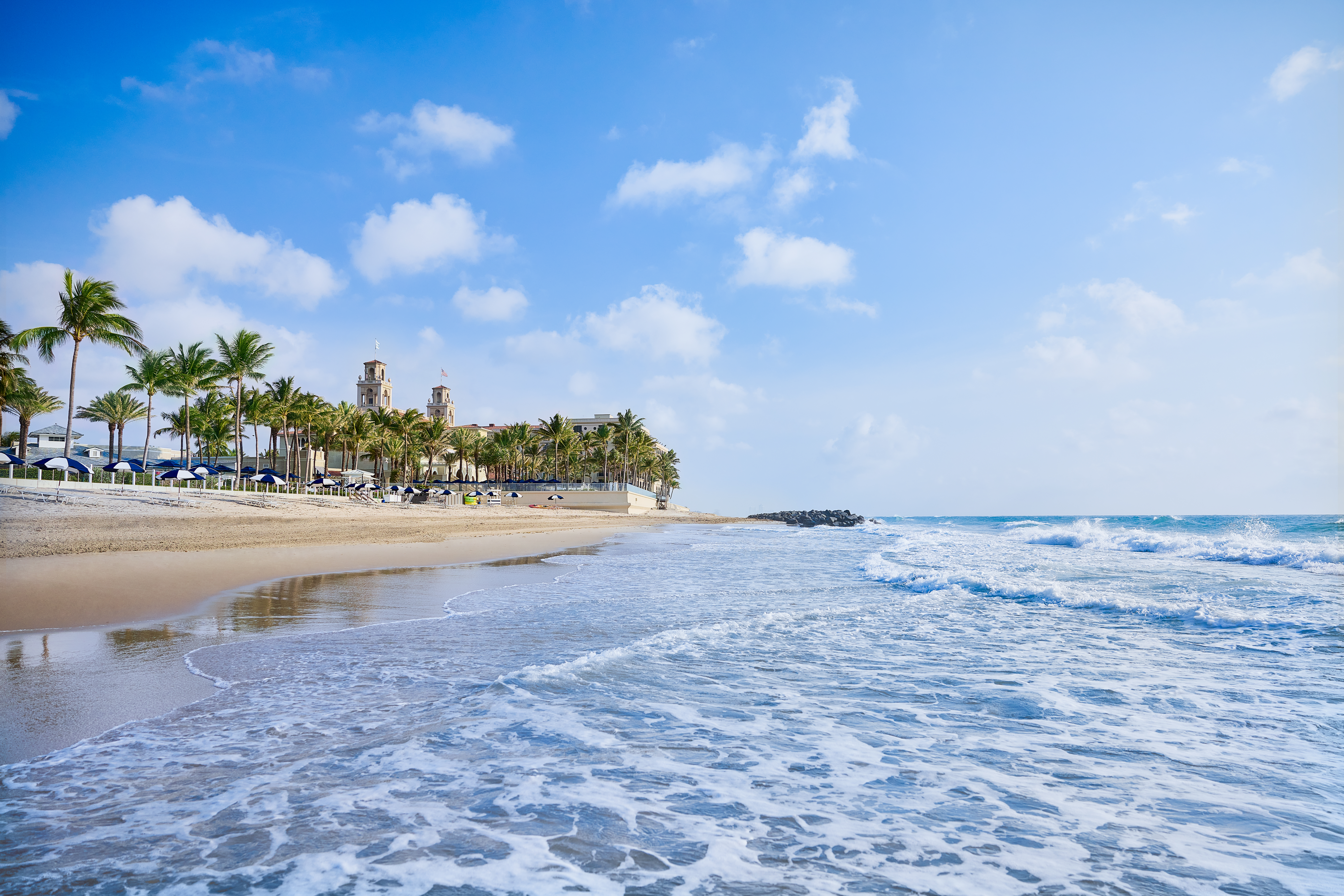 Tropical beach with palm trees, resort buildings, and waves rolling onto sandy shore under blue sky.