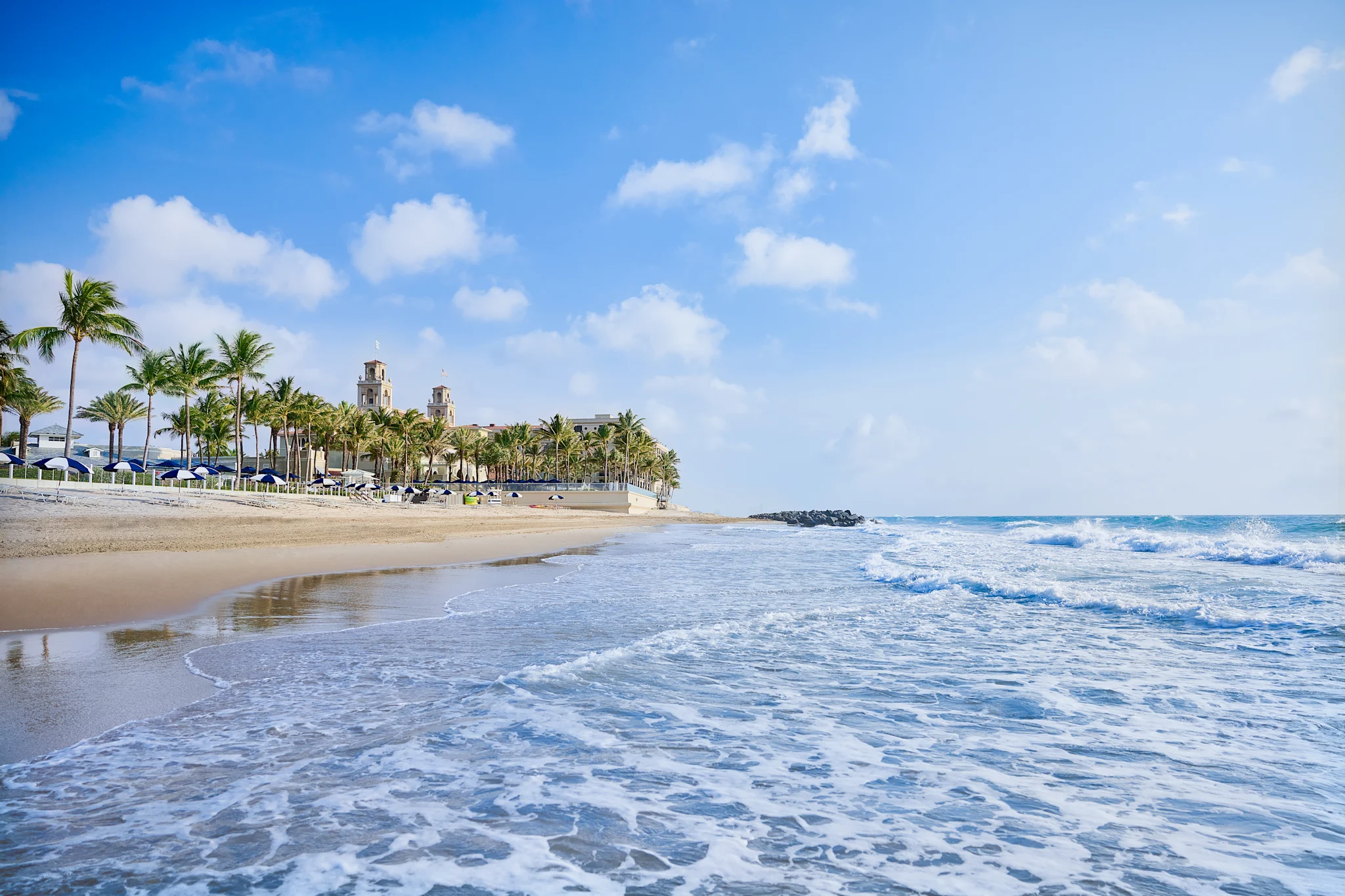 Tropical beach with palm trees, resort buildings, and waves rolling onto sandy shore under blue sky.