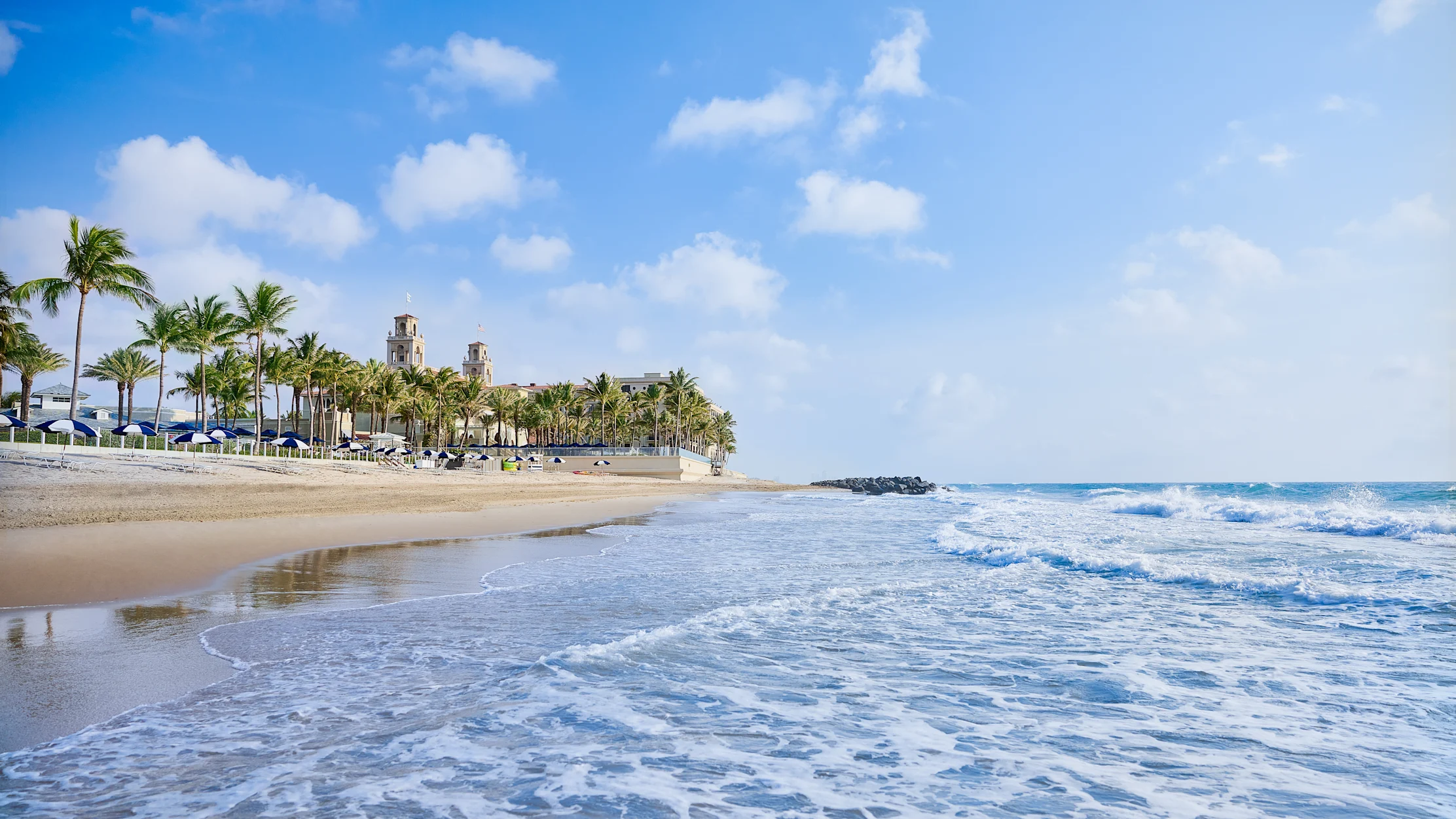 Tropical beach with palm trees, resort buildings, and waves rolling onto sandy shore under blue sky.