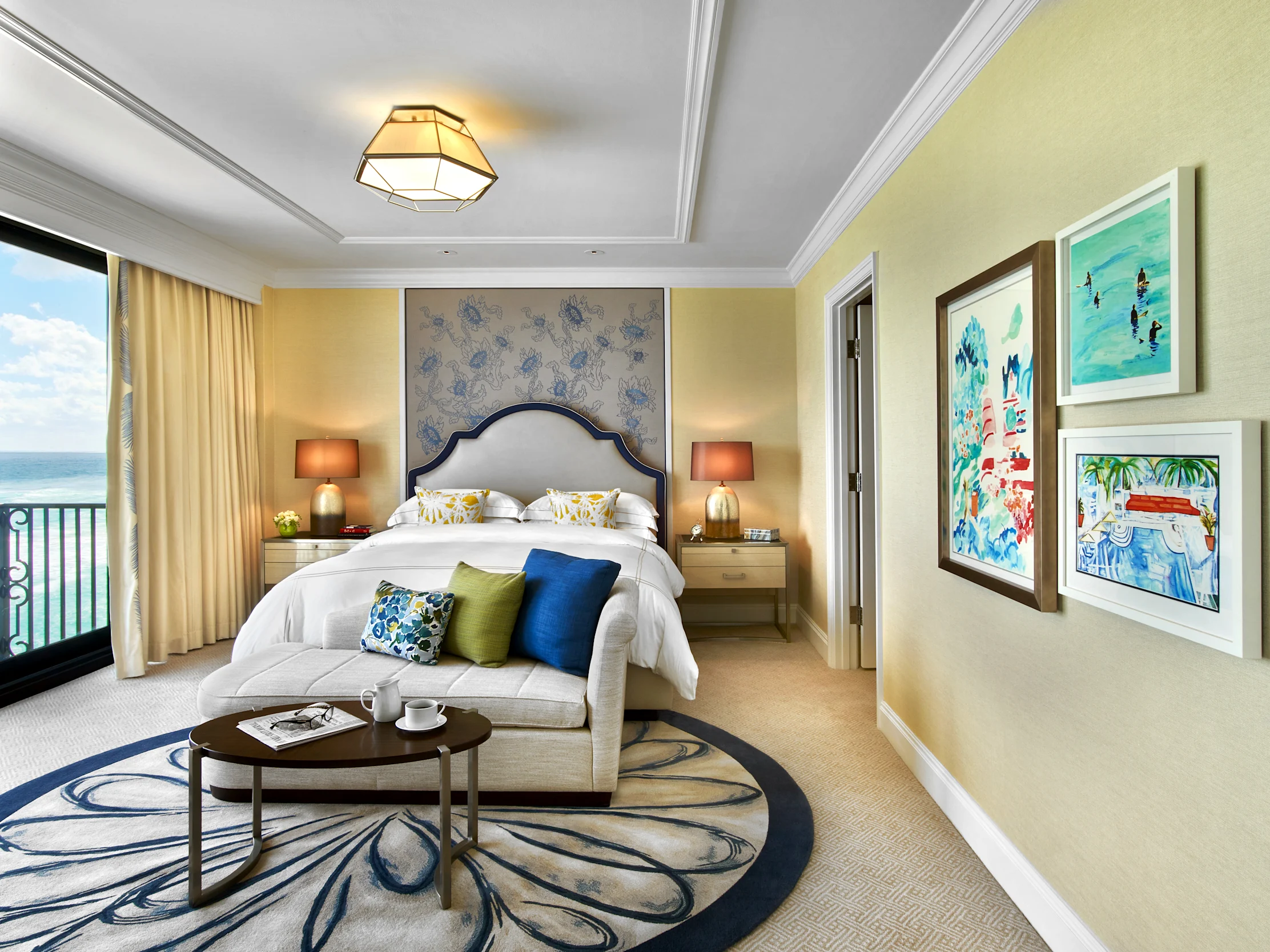 Elegant hotel bedroom with ocean view, upholstered headboard, seating area, and coastal artwork.