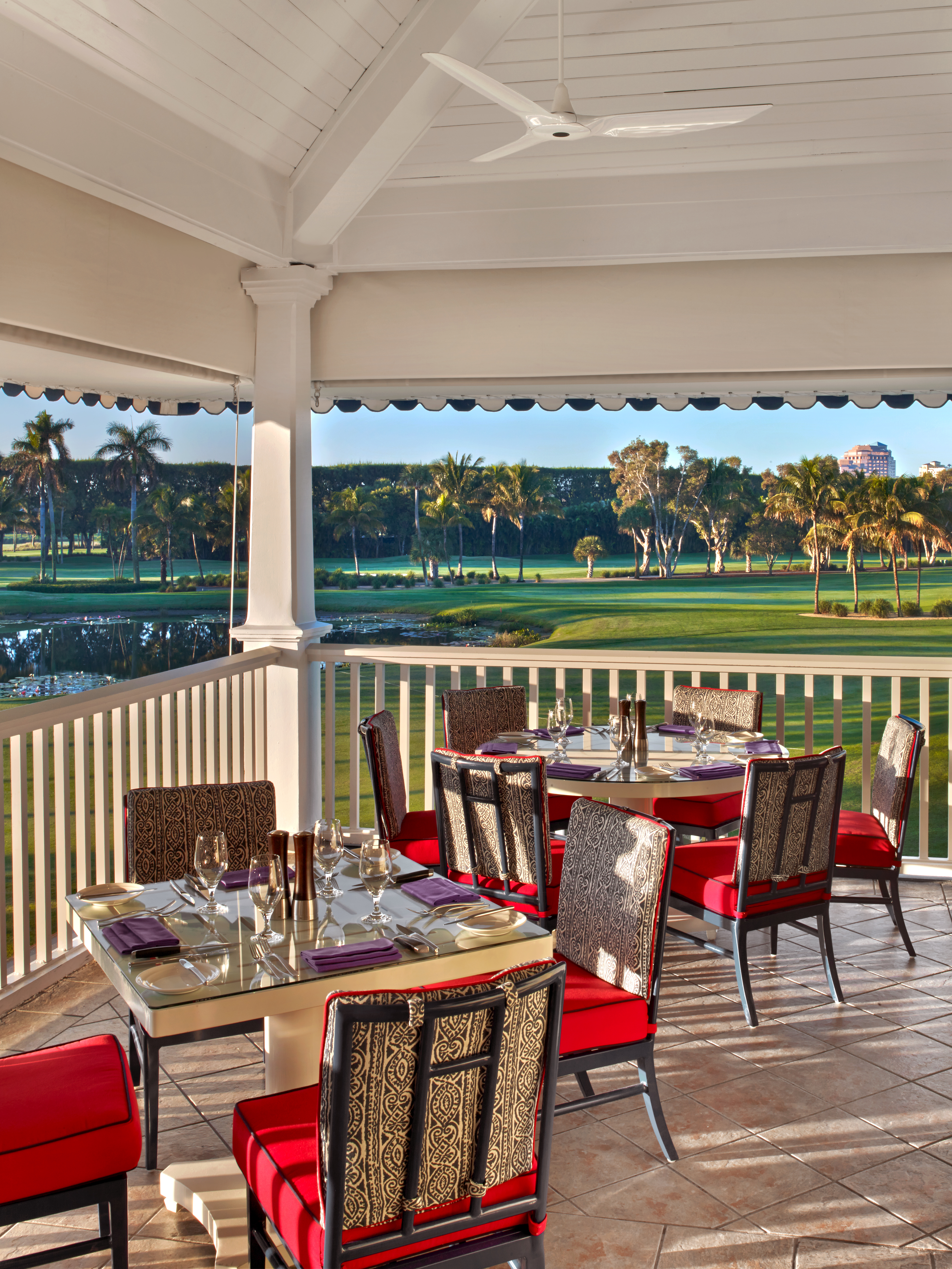 Covered patio dining area with red-cushioned chairs overlooking a lush golf course and palm trees.